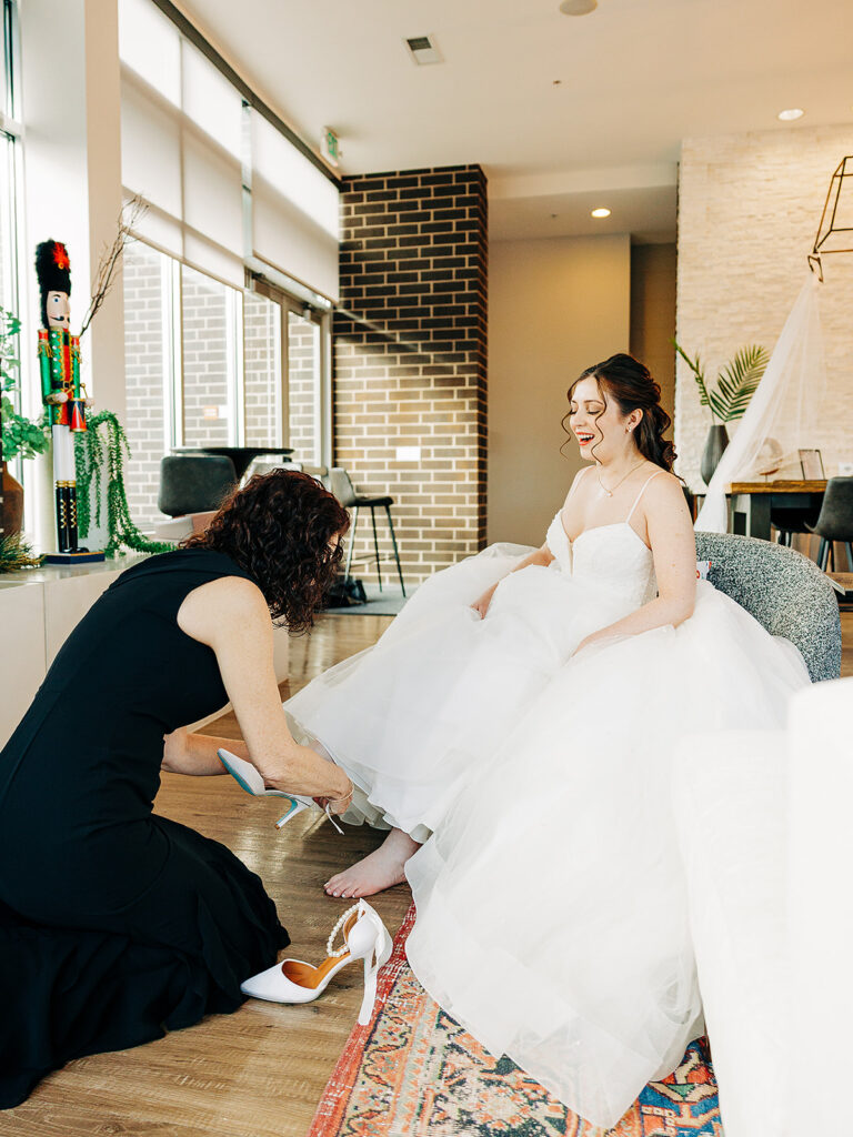 A bride in a white wedding dress sits on a chair and smiles as another woman in a black dress kneels to help her put on a high-heeled shoe in a sunlit modern room.