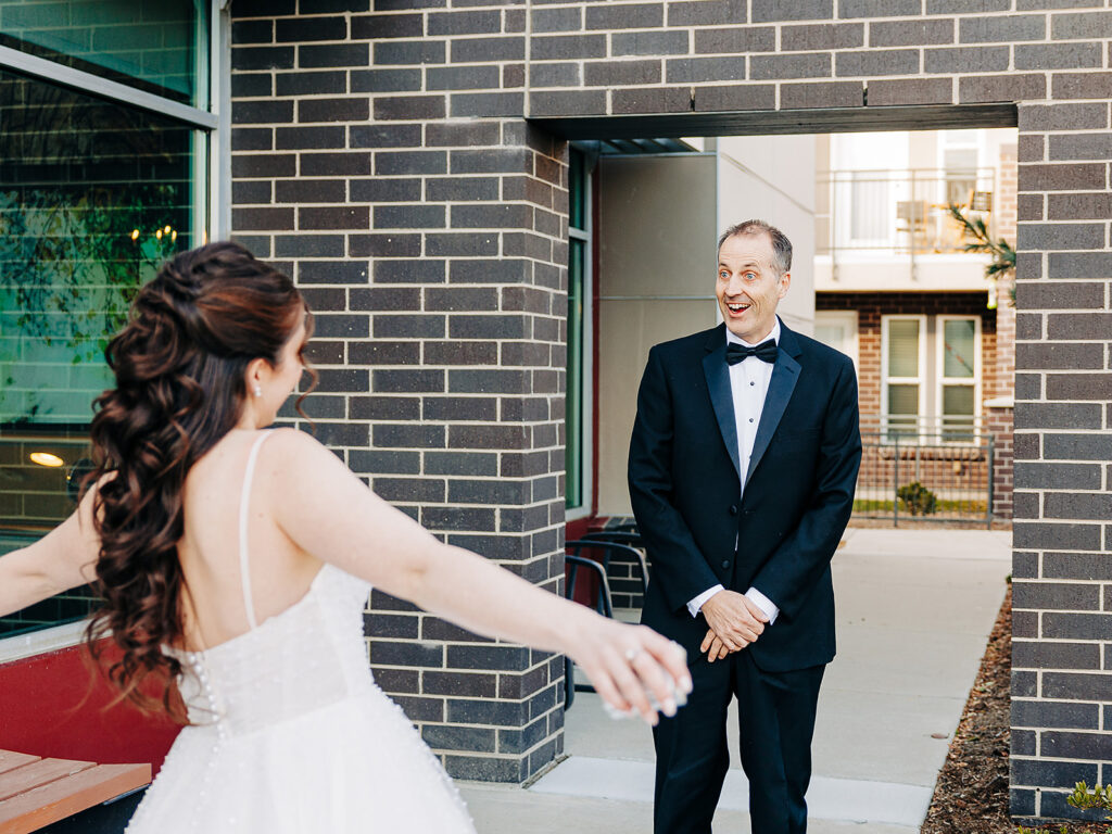 A bride in a white wedding dress approaches a smiling man in a black tuxedo, standing outside a brick building. The man looks pleasantly surprised as the bride holds her arms open wide.