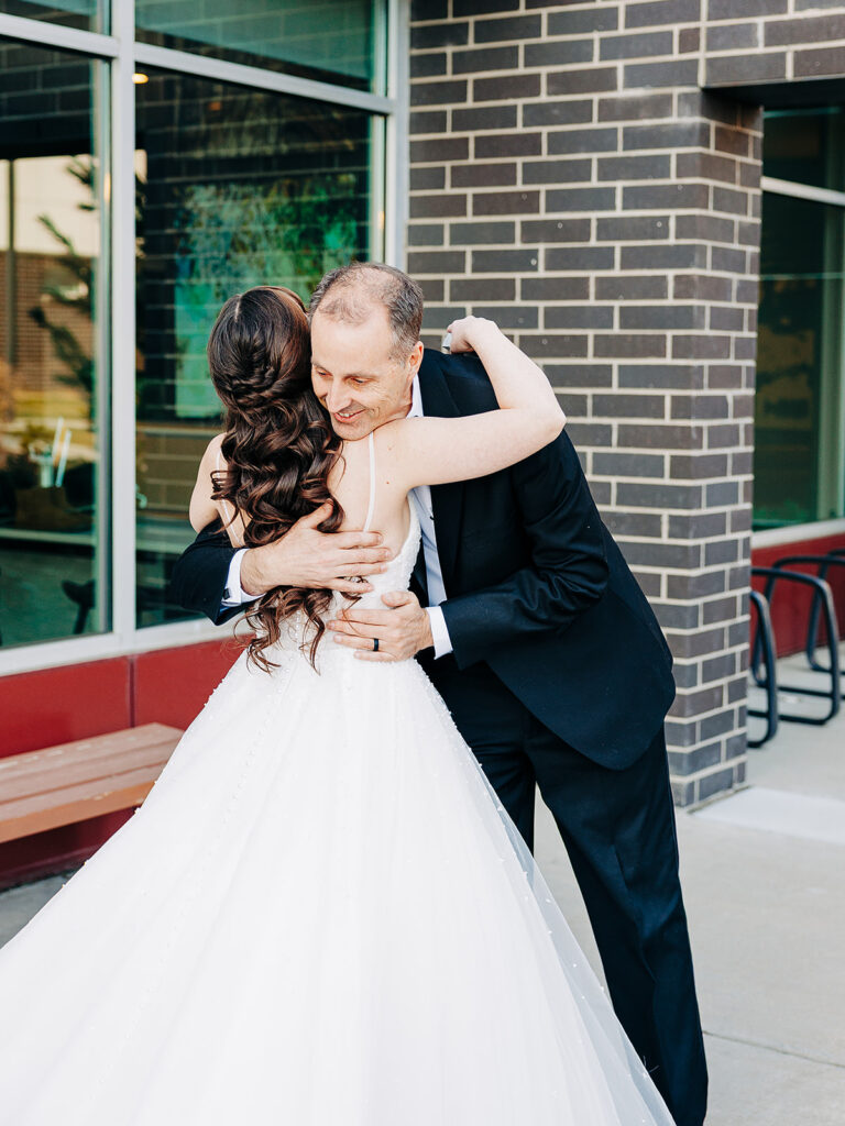 A bride in a white gown with long, wavy hair hugs a man in a black suit outside a brick building with large windows. The scene appears emotional and joyful.