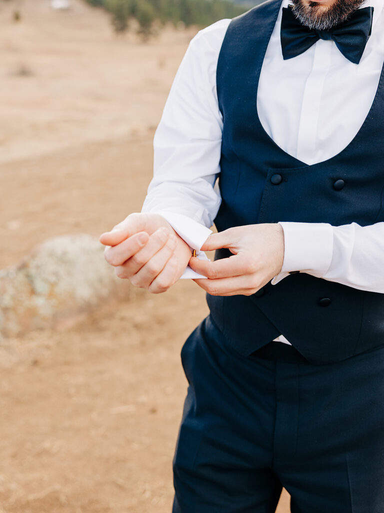 A man dressed in a formal navy blue suit with a black bow tie fastens his cufflink outdoors in a dry, brown landscape. The image focuses on his torso and hands, not showing his face.