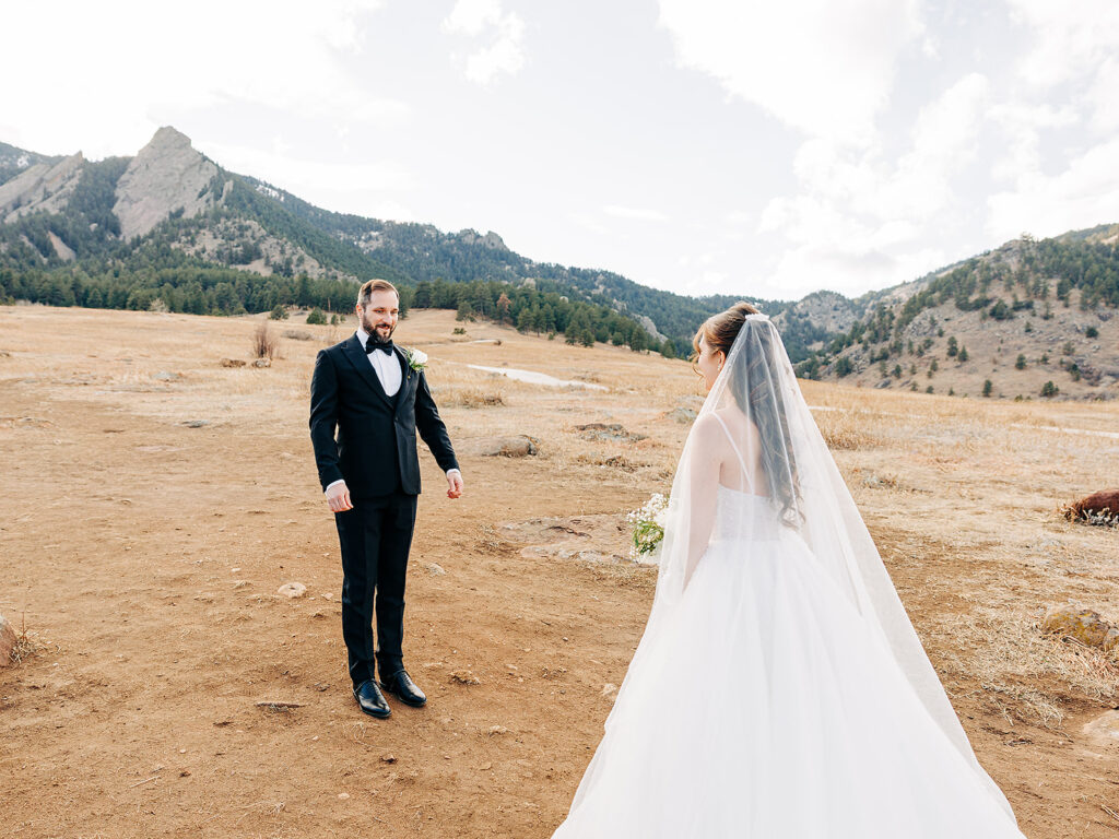 A bride in a white gown and veil approaches a groom in a black tuxedo, both standing outdoors on a dry, open field with mountains and trees in the background under a partly cloudy sky.