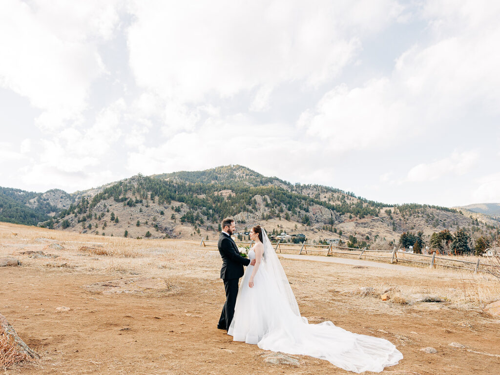 A bride and groom stand together outdoors in a dry, open field with mountains and a cloudy sky in the background. The bride wears a white gown, and the groom is dressed in a black suit.