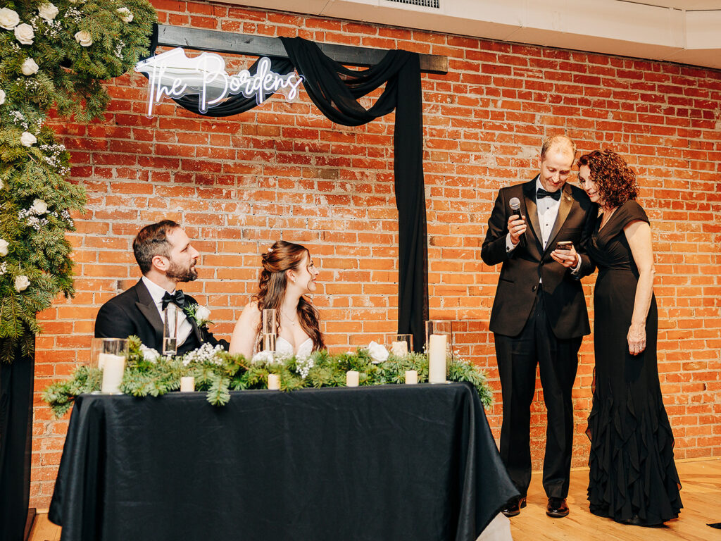 A bride and groom sit at a table decorated with greenery and candles, smiling at two people in formal attire who are giving a toast with a microphone. A neon sign reading The Bordens hangs on a brick wall behind them.