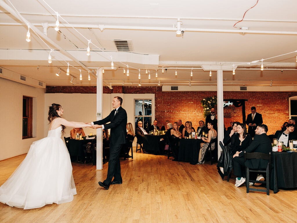 A bride and groom share their first dance in a warmly lit reception hall as guests seated at tables watch and smile.