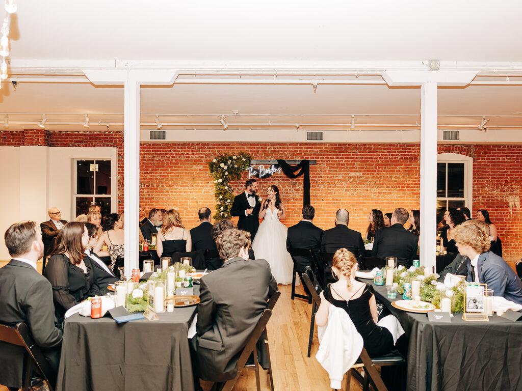 A bride and groom stand together giving a speech at their wedding reception, facing seated guests at decorated tables in a bright room with brick walls and wooden floors.