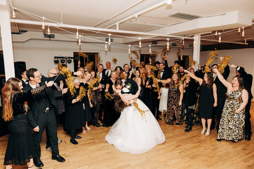 A bride and groom kiss in the center of a crowded dance floor as guests cheer and wave gold streamers. Everyone is dressed formally, smiling and celebrating in a warmly lit indoor venue.