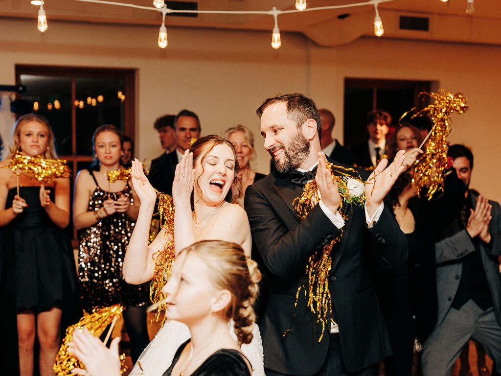 A bride and groom in formal attire smile and clap joyfully, surrounded by guests holding gold tinsel pom-poms at a festive indoor celebration with string lights.