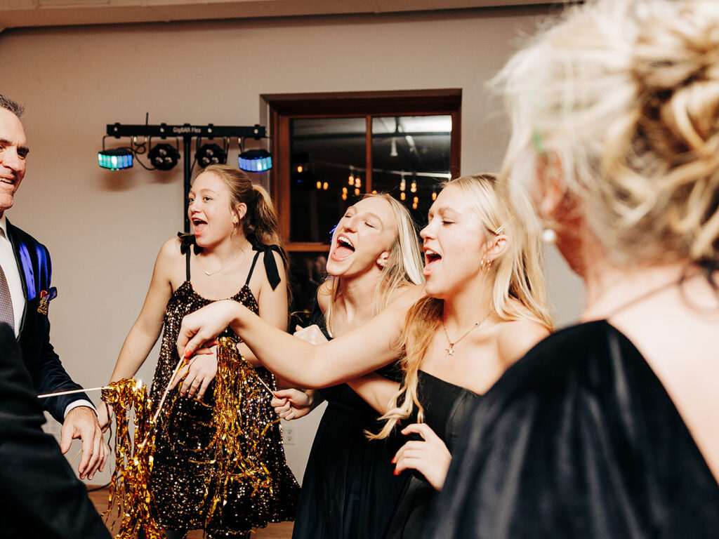 Four young women in black dresses, laughing and singing as they dance at a party, with one holding gold pom-poms. Warm indoor lighting and a man in a suit are visible in the background.