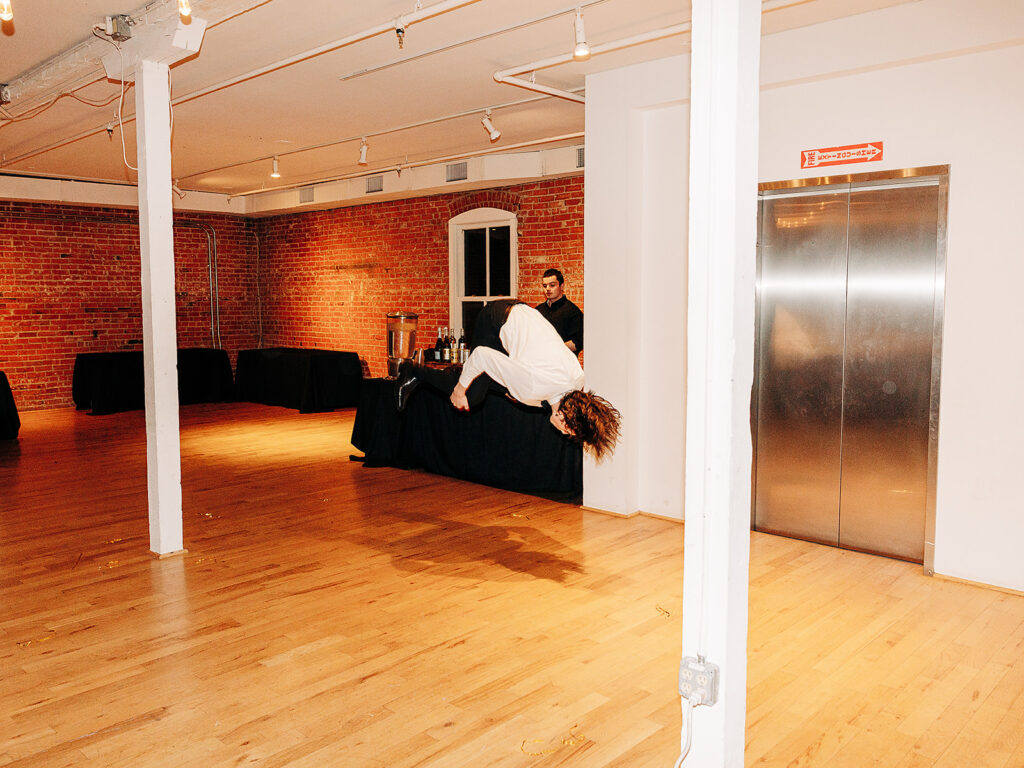 A person in white clothes is caught mid-air as they tumble or fall backward over a black-covered table in a spacious room with brick walls, wooden floors, and an elevator. Another person stands behind the table, watching.