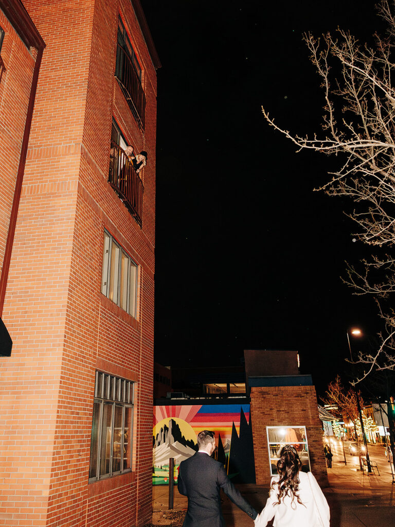 A couple stands outside at night, looking up at a person waving from a balcony of a tall red brick building. Colorful mural and bare tree branches are visible in the background.