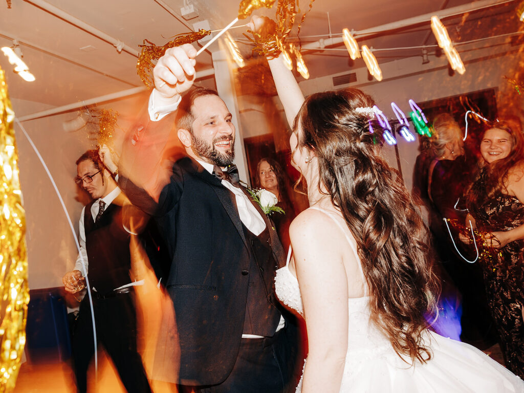 A bride and groom dance joyfully at their wedding reception, surrounded by guests, golden streamers, and colorful party lights, creating a lively and festive atmosphere.
