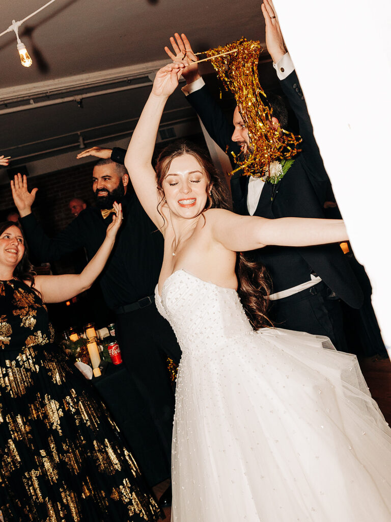 A bride in a strapless white gown dances joyfully with guests, arms outstretched, eyes closed, and smiling. Partygoers around her hold gold streamers and celebrate enthusiastically indoors.