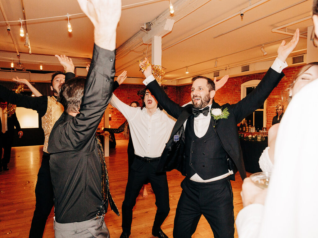 A group of men in formal attire energetically dance with arms raised at an indoor celebration, smiling and enjoying themselves in a warmly lit room with exposed brick walls and wooden floors.