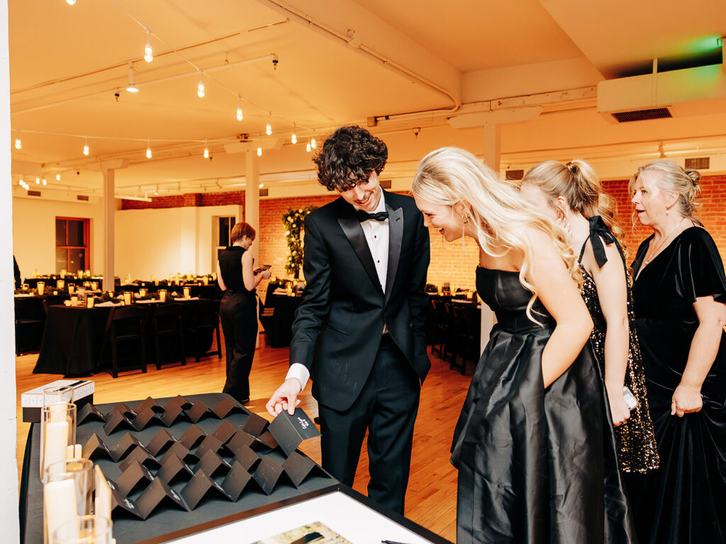 A group of elegantly dressed people in formal evening wear smile and examine black envelopes arranged on a table at a brightly lit indoor event. The background features decorated tables and warm lighting.