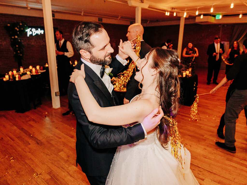 A bride and groom dance together on a wooden floor at their wedding reception, smiling at each other. Guests and candlelit tables with decorations are visible in the warmly lit background.