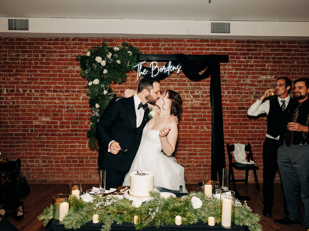A bride and groom share a kiss behind their wedding cake at a decorated reception table. Guests in formal attire celebrate nearby. A neon sign reading The Boghers hangs on a brick wall with greenery and white flowers.