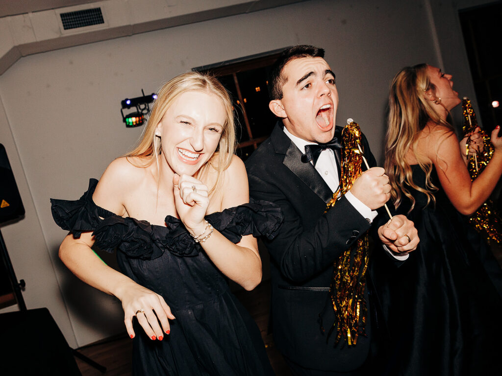 Two people dressed in formal black attire laugh and cheer while holding gold tinsel pom-poms at an indoor event, with lively expressions and another person partially visible in the background.