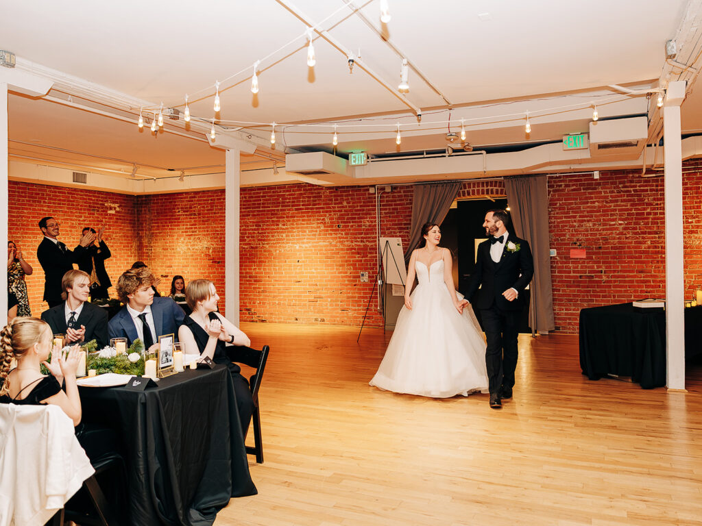 A bride and groom smile as they enter a reception hall with exposed brick walls. Seated guests watch and take photos. The room has string lights and black tablecloths, creating a warm, festive atmosphere.