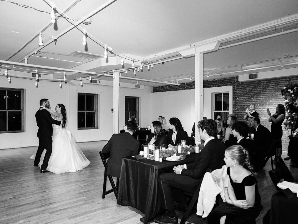 A bride and groom share their first dance in a bright, modern venue as guests seated at black-clothed tables watch and smile. String lights hang from the ceiling, and the room is decorated simply with flowers.