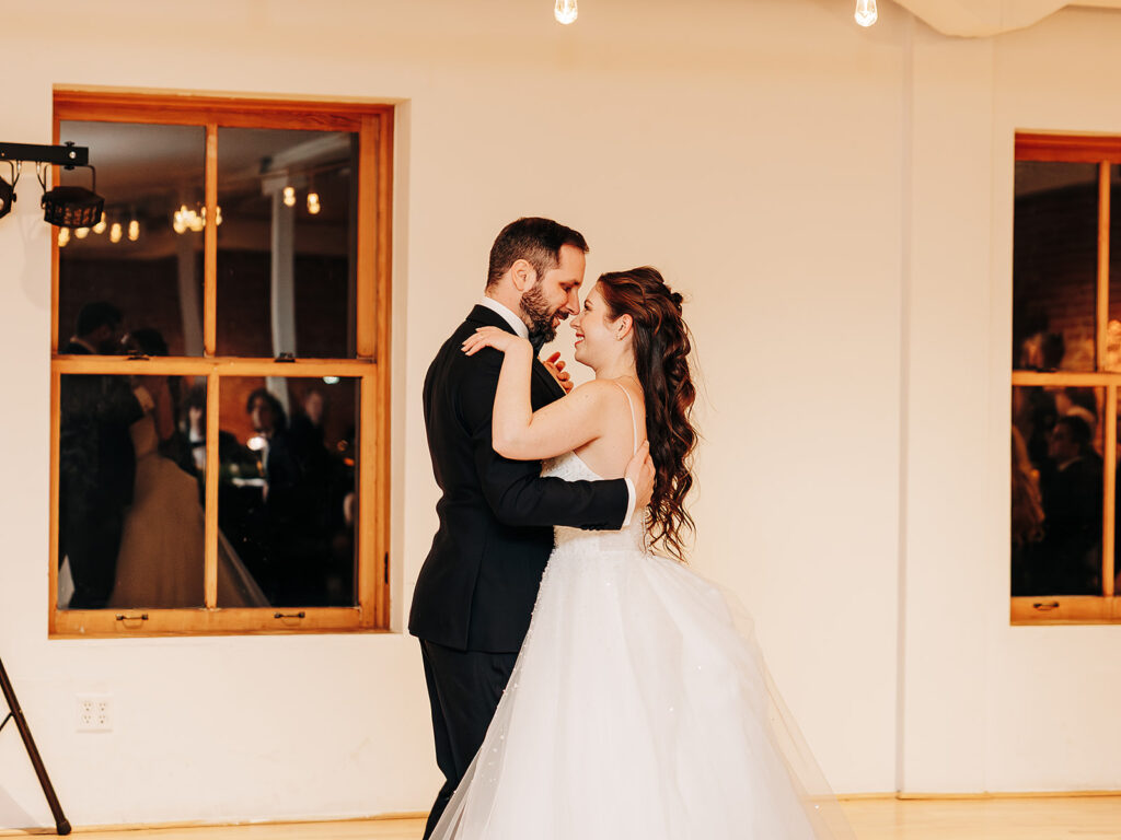 A bride in a white gown and a groom in a black suit share their first dance in a bright, warmly lit room with large windows reflecting wedding guests.