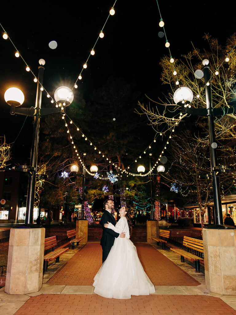 A bride and groom stand together under string lights in an outdoor plaza at night, surrounded by benches and trees decorated with holiday lights. They gaze at each other, smiling, in the center of a brick pathway.