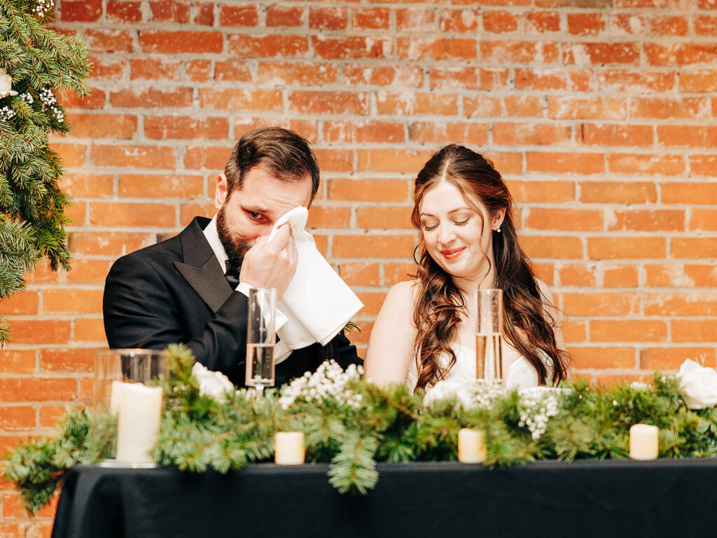 A groom wipes his face with a napkin while sitting next to his smiling bride at a table decorated with greenery and candles, against a brick wall backdrop.