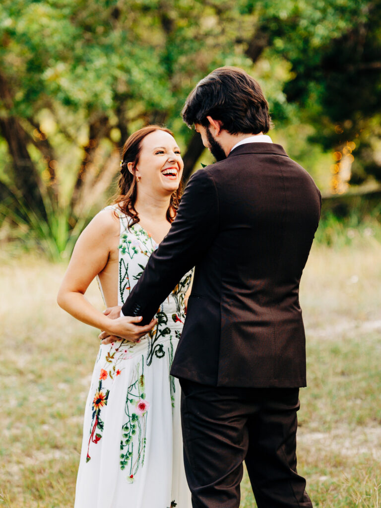 A woman in a white floral dress and a man in a dark suit stand outdoors, holding hands and smiling at each other in a grassy, tree-filled setting. The woman is laughing joyfully.