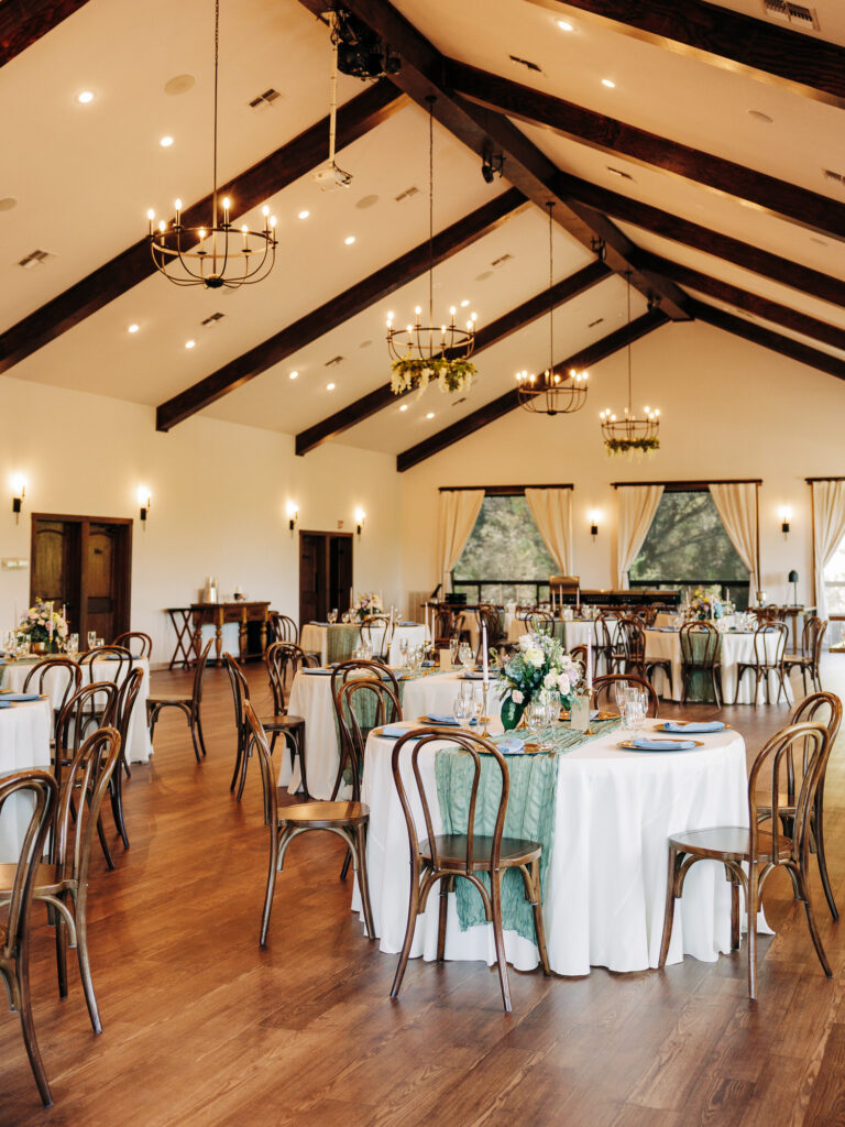 Elegant banquet hall with wooden floors, round tables covered in white cloths, green table runners, and floral centerpieces. Wooden chairs surround the tables, and large windows with curtains let in natural light. Chandeliers hang from ceiling.