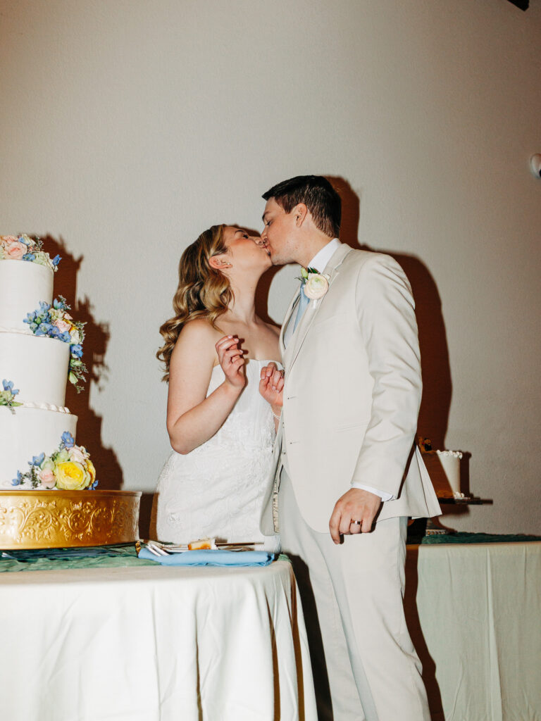 A bride and groom in wedding attire kiss while standing next to a three-tiered white wedding cake decorated with flowers. The bride holds a fork and plate, suggesting they’ve just cut the cake.