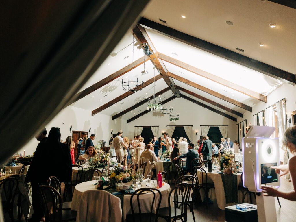 A lively indoor wedding reception with guests mingling around decorated tables. The room features wooden beams, floral centerpieces, and a photo booth with a bright ring light in the foreground.