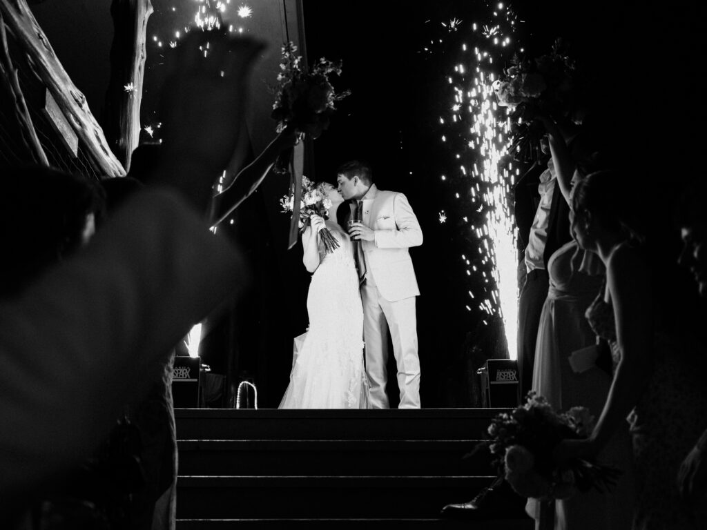 A bride and groom kiss on a staircase at night, surrounded by friends and family, with fireworks sparkling behind them in celebration. The image is in black and white.