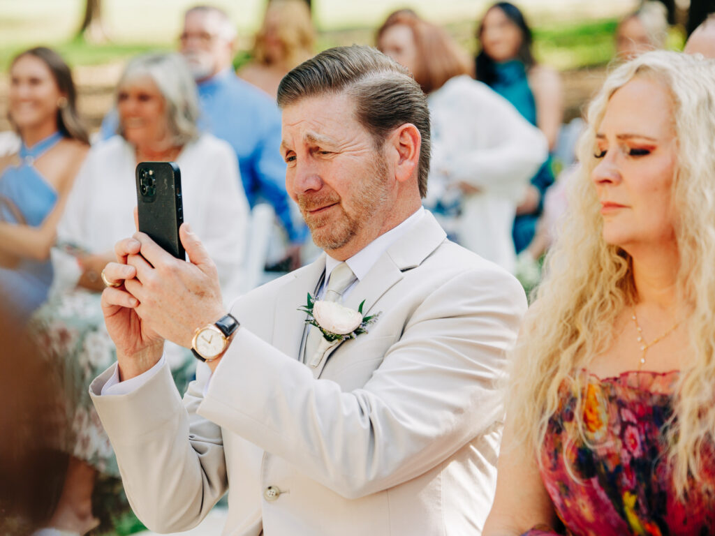 A man in a light suit takes a photo with his phone at an outdoor event, possibly a wedding. He wears a boutonniere and watch, while a woman with long blonde hair in a colorful dress sits beside him. Other guests are seated behind them.