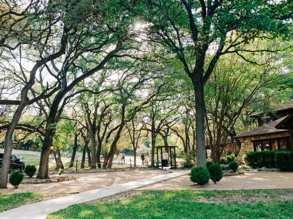 A sunlit park with tall, leafy trees, a paved walkway, and a rustic wooden building. A few people walk in the distance, and green grass and bushes line the foreground.