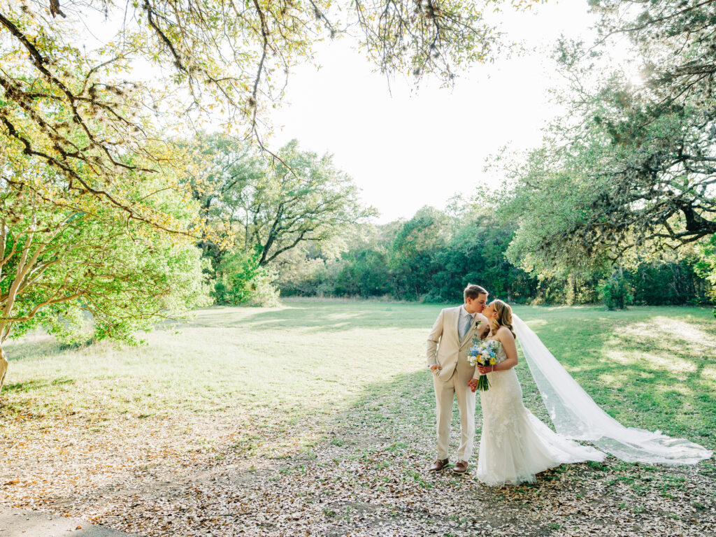A bride and groom stand together outdoors under trees, the groom in a light suit and the bride in a white dress with a long veil, holding a bouquet and sharing a kiss in the sunlight.