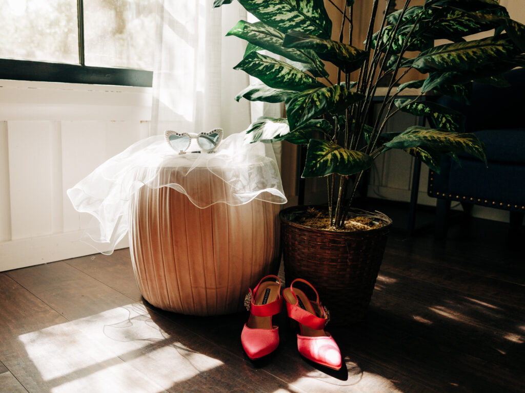 A pair of red shoes, a white veil, and sparkly sunglasses are arranged on and beside a round ottoman next to a large potted plant in a sunlit room with wooden floors.