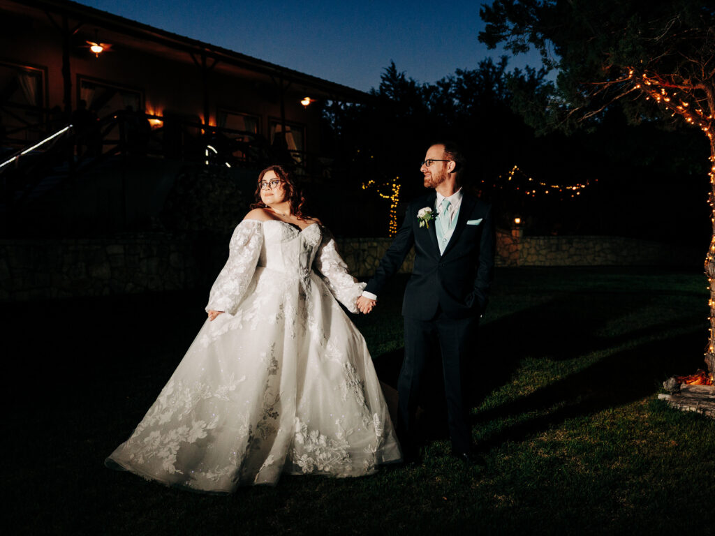 A bride in a white lace gown and a groom in a dark suit hold hands outdoors at night, illuminated by string lights and a tree wrapped in lights, with a building in the background.