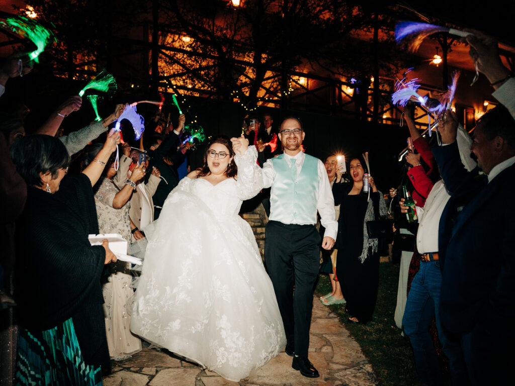 A bride in a white gown and groom in a vest walk hand-in-hand down an outdoor path, smiling, as guests cheer and wave glow sticks and streamers in a festive nighttime celebration.