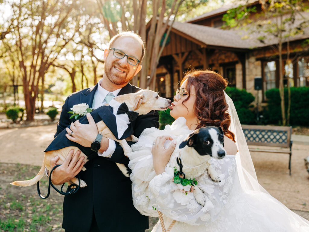 A bride and groom smile outdoors, each holding a dog dressed in wedding attire. The groom wears a suit and holds a tan dog in a tiny tuxedo, while the bride in a white dress kisses a black and white dog in a frilly outfit.
