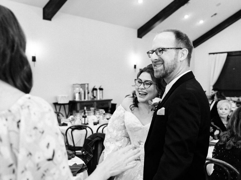 A smiling bride and groom stand together at their wedding reception, greeting a guest. The bride wears glasses and a white dress; the groom wears glasses and a suit. Guests sit at round tables in the background.