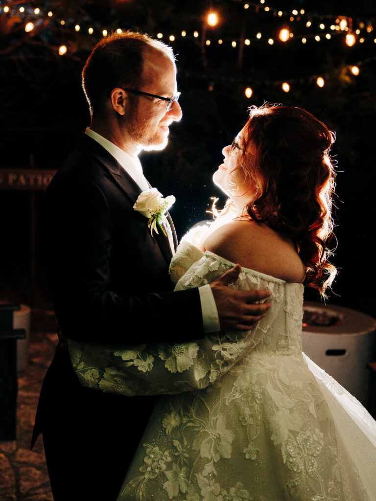 A bride and groom in formal attire embrace under string lights at night, gazing at each other and smiling. The bride wears a white floral gown, and the groom wears a dark suit with a boutonniere. Warm lights create a romantic atmosphere.