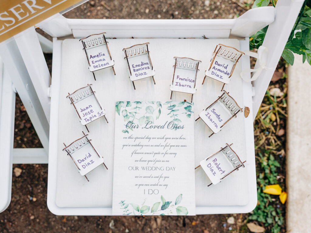 Eight small wooden chairs with name cards are arranged around a printed sign on a white chair outdoors. The sign reads “Our Loved Ones” and mentions honoring loved ones at a wedding.