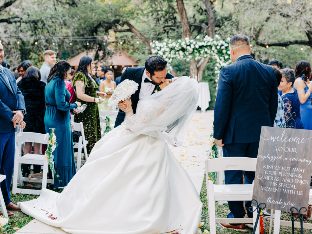 A bride and groom share a kiss at their outdoor wedding ceremony, surrounded by guests. The groom dips the bride, who holds a bouquet and wears a long white gown. A sign asks guests to put away phones and cameras for the moment.