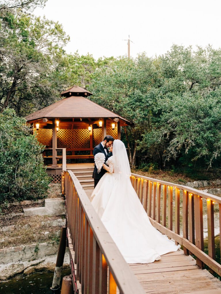 A bride and groom stand close together on a wooden bridge with string lights, facing each other, with a gazebo and lush green trees in the background.