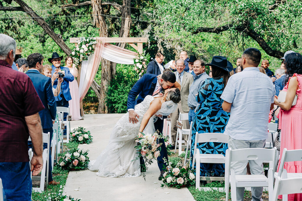 A bride and groom share a kiss at the end of an outdoor aisle at Scenic Springs Helotes, surrounded by standing guests. Floral arrangements and a wooden arch decorate the background, with trees and greenery completing the celebratory wedding scene.