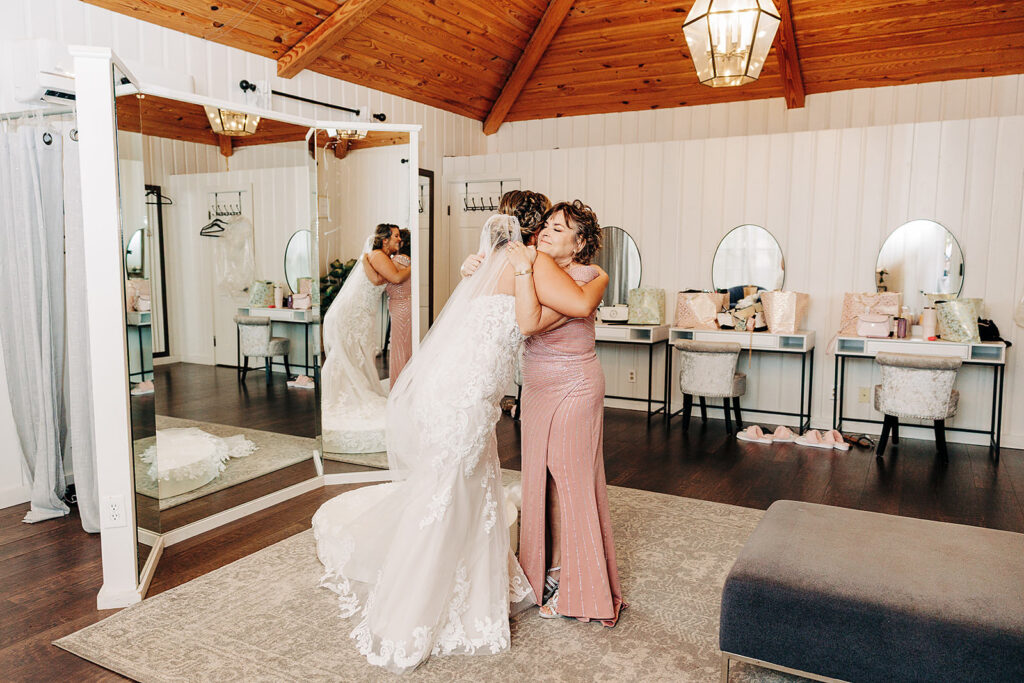A bride in a white gown and veil hugs a woman in a pink dress inside the bright, wood-ceilinged dressing room at Scenic Springs Helotes, with mirrors, vanity tables, and wedding accessories visible in the background.