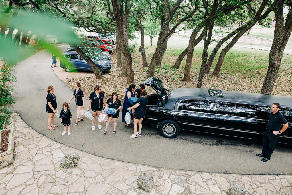 A group of people, mostly women and children, stand near a black limousine parked on a tree-lined driveway at Scenic Springs Helotes. Some are holding bags and a cooler, while a man stands by the limo and several cars are parked in the background.