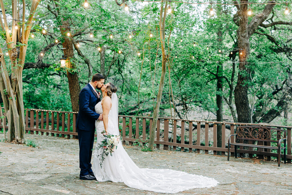 A bride and groom stand close together, embracing on a stone patio under string lights at Scenic Springs Helotes, surrounded by green trees. The bride wears a long white gown and veil, while the groom wears a navy suit.