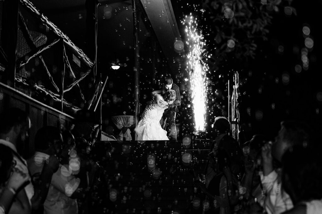 A bride and groom share a romantic dance at night at Scenic Springs Helotes, dipped in an embrace on a staircase, surrounded by guests and dramatic sparks, with bubbles floating in the foreground.