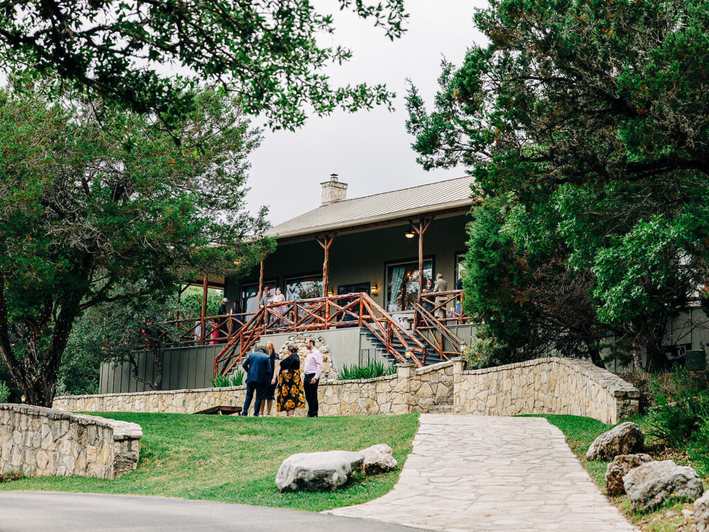 A group of people gather outside a rustic building with a wooden porch and stone walkway, surrounded by greenery and trees on a cloudy day.