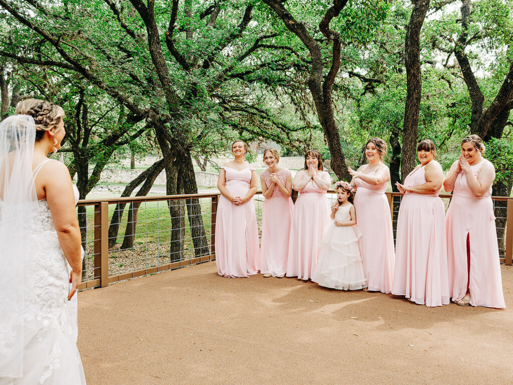 A bride in a lace gown and veil stands facing six bridesmaids and a flower girl in pale pink dresses, who smile and clap with delight outdoors on a tree-lined patio.